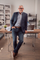 Portrait of a smiling senior manager leaning on his desk in a modern office, wearing a blue jacket...
