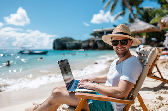 Sea waves gently crashing against barefoot young person seated, typing on a laptop outdoors