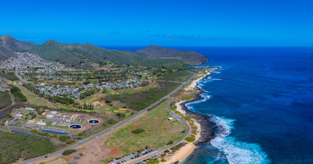 Fototapeta premium Aerial view of Oahu, Hawaii, showing a highway along the coast, lush greenery, a small beach, water treatment structures, and a residential area by mountains.