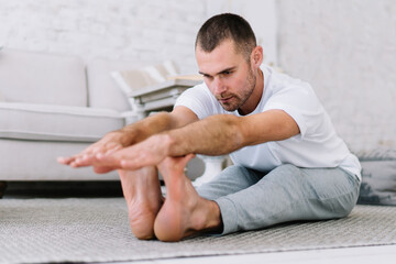 Fototapeta premium Young caucasian male in casual wear bending stretching neck muscles during morning workout, positive man enjoying free time and yoga training sitting in asana keeping healthy lifestyle