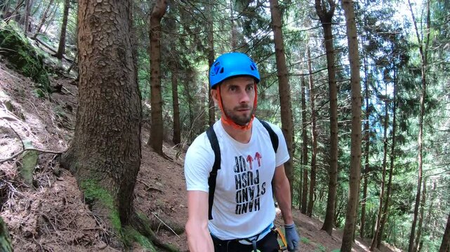  An adventurer on the Via Ferrata Corna delle Capre pauses, his blue helmet a bright spot against the backdrop of the dense, green woodland.