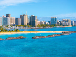 Naklejka premium View of Honolulu's skyline with high rise buildings, turquoise ocean waters, sandy beach, palm trees, rock formations, and a marina in the distance.