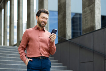 Successful man walking through the city outside an office building with a phone in his hands. Businessman using an app on his smartphone, typing messages, and browsing social media, smiling.