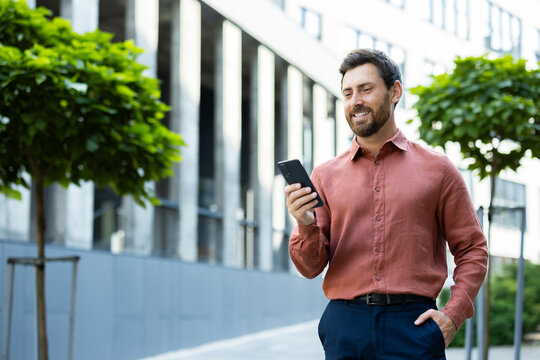 Successful man walking through the city outside an office building with a phone in his hands. Businessman using an app on his smartphone, typing messages, and browsing social media, smiling.