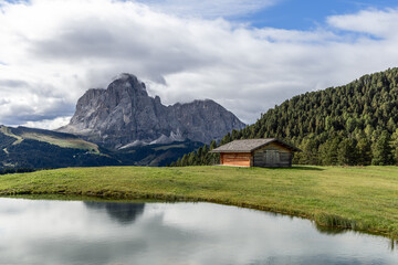 The peaceful alpine landscape features a small pond, a rustic cabin, and the dramatic Sassolungo peak under a partly cloudy sky in the Dolomites