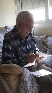 older man on sofa reviewing paper invoice, blue checked shirt and protective fabric armchair, in the background leaning on canes