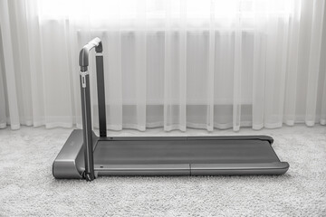 A modern, compact treadmill with a foldable handlebar is placed on a soft carpet in a bright,...