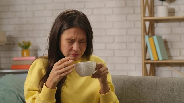 Young woman spending day at home sitting on the sofa in the living room, holding cup and drinking tea, disgusting taste of drink, upset negative expression.