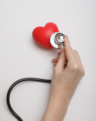 Hand holding stethoscope checking red heart on white background, symbolizing cardiology, healthcare, heart disease prevention, and medical diagnostics. Concept of heart health, love, and wellness.