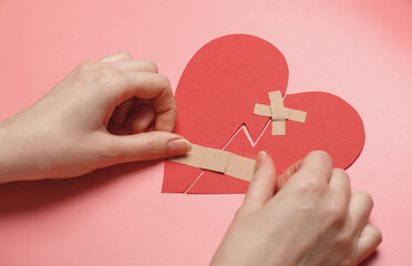 Hands carefully applying band-aids to broken red paper heart on pink background, symbolizing healing, love recovery, emotional repair. Touching concept for heartbreak, forgiveness, relationships
