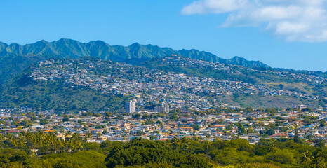 Aerial view of a hillside residential area on Oahu island, Hawaii, with lush green mountains, dense housing, palm trees, and a clear blue sky.
