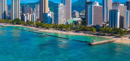 Turquoise waters meet sandy shores at Waikiki Beach, with a pier, palm trees, high rise hotels, and green mountains in the background.