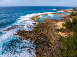 Aerial view of Oahu's rocky coastline with turquoise waters, sandy beach, lush green vegetation, and waves crashing against rugged rock formations.