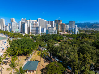 Aerial view of Honolulu, Oahu, Hawaii, featuring Waikiki Beach, a park with palm trees, modern high rises, a clear blue sky, and distant mountains.