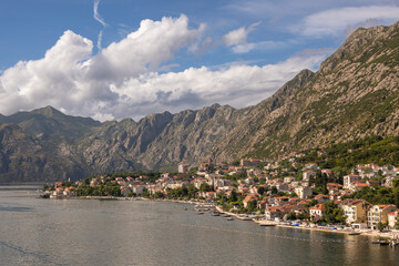Fototapeta premium Kotor, Montenegro - July 2, 2024: NW side of the city landscape along bay shoreline with gray rocky mountain range in back under blue cloudscape. Short piers and small boats