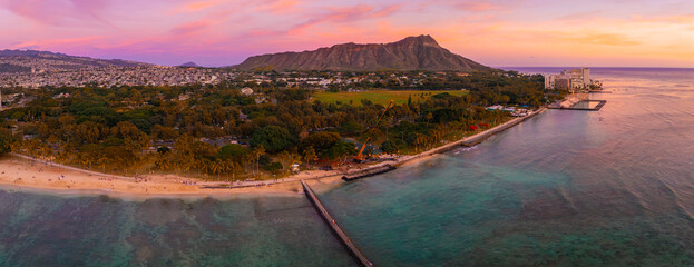 Obraz premium Aerial view of Waikiki Beach in Honolulu, Hawaii, with Diamond Head Crater in the background, sandy shores, turquoise waters, and urban structures at sunset.