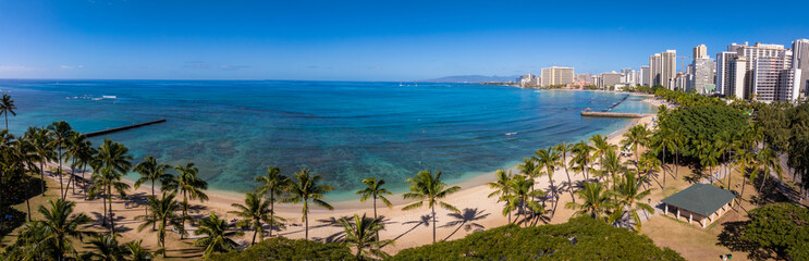 Aerial view of Waikiki Beach in Honolulu, Oahu, Hawaii, featuring turquoise waters, sandy shores, palm trees, piers, and high rise buildings under a clear sky.