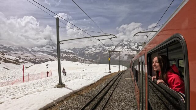 Happy tourist woman smiling and leaning out of red train window, enjoying stunning winter mountain views in the Swiss Alps. Perfect for travel, adventure, and lifestyle themes