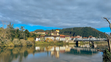 A peaceful riverside village with vibrant houses reflecting on the calm water. Surrounded by lush mountains under a dramatic sky, evoking tranquility and timeless beauty.