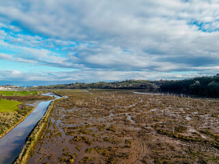 An expansive aerial view of a marshland featuring a winding river, surrounded by green hills and forests. The dramatic cloudy sky adds depth to this tranquil natural landscape.