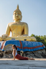 A woman gracefully performs yoga in front of a majestic golden Buddha statue, embracing tranquility...