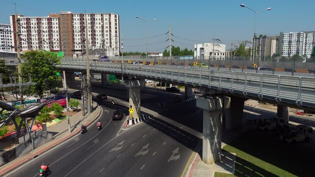 Aerial view showcasing modern urban transportation in a bustling city. An elevated train moves along its track while cars and motorcycles navigate the circular intersection below. The scene captures t