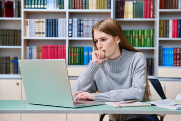 Sad teenage student girl preparing in classroom library looking at laptop