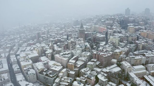 A drone hovers over snow-covered Istanbul, revealing the Galata Tower and surrounding historical structures. As snowflakes continue to fall, the city takes on the enchanting atmosphere of winter.