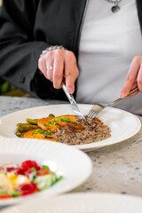 A close-up of a person using a knife and fork to cut into a plate of delicious food, including meat, vegetables, and a creamy sauce, set against a soft-focus background.