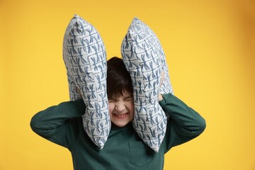 Annoyed boy covering his ears with pillows due to loud sound on yellow background