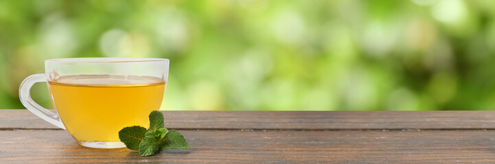 Aromatic mint tea in glass cup and fresh leaves on wooden table outdoors. Banner design with space...