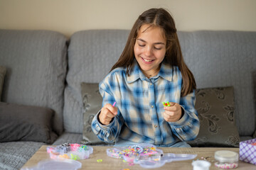Smiling young girl making diy loom band bracelets at home, sitting on the sofa and concentrating on...