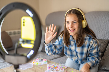 Smiling preteen girl wearing headphones, waving at the camera while recording a diy bracelet-making...