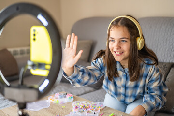 Smiling young girl with headphones waving, broadcasting a diy craft tutorial using a smartphone and...