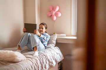 Happy young girl is using a digital tablet while sitting comfortably on her bed in a cozy bedroom,...