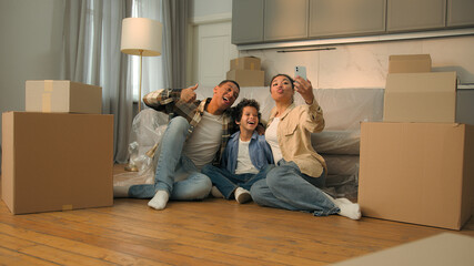 Naklejka premium Happy African American family on floor at home moving day relocation smile joyful parents little son child kid boy posing selfie photo mobile phone grimacing have fun weekend buy rent house mortgage