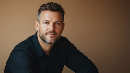 Obraz premium Attractive man in his late thirties in black shirt with short hair and beard looking at camera and smiling sitting on beige background.