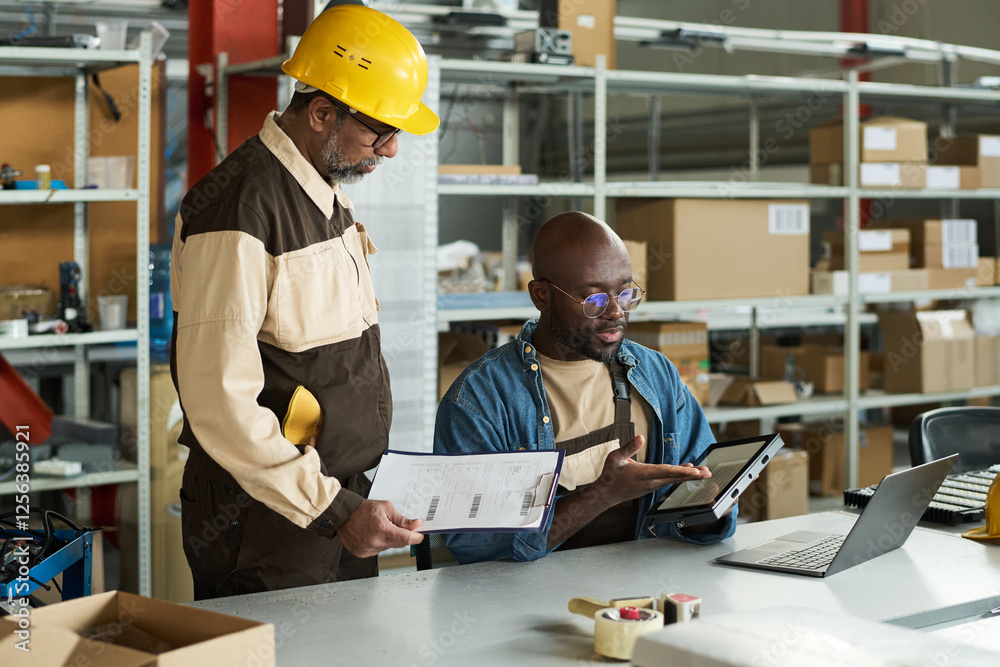 Poster young male worker pointing at lamp while sitting by workplace and explaining his boss principles of 
