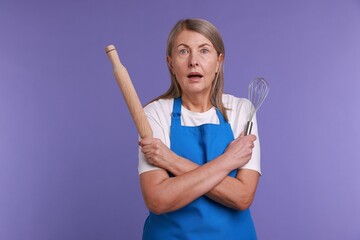 Surprised woman with rolling pin and whisk on violet background
