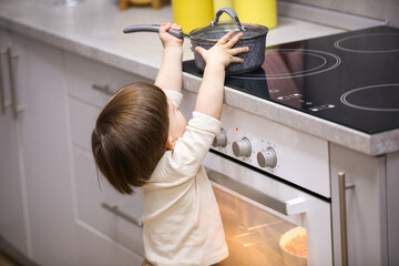 Little boy playing with pot on stove in kitchen, back view. Dangerous situation