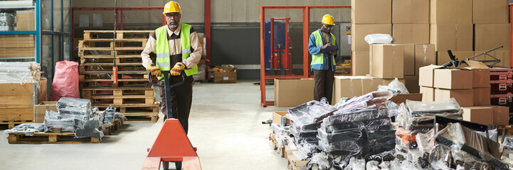 Header of mature engineer in workwear pushing empty pallet truck while moving along stacks of packed items in warehouse or plant