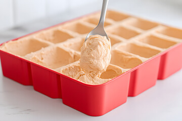 Frozen Dessert Preparation: A spoon scoops creamy mixture from a red mold, ready to freeze into individual portions on a white marble surface.