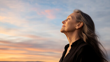 Woman gazes at the sunset with a serene expression, showcasing natural aging and contemplation against a pastel sky.