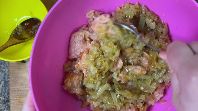 Mixing raw minced chicken and fried onions with a spoon in a bowl. The process of making chicken frikadelles. Top view.