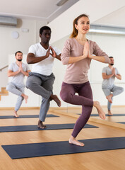 Obraz premium Full-length portrait of positive fit young woman standing on one leg in Eka Pada Utkatasana pose with clasped hands in namaste gesture during group yoga training in gym