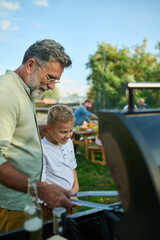 Grandfather teaching grandson how to grill at family barbecue