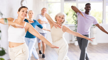 Obraz premium In fitness studio, youthful female coach is leading class for seniors. Instructor is teaching older students fundamental steps of hip hop dance in dance lesson