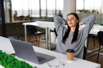 Fototapeta premium Young business woman smiling and relaxing in a bright office, enjoying a break at work with hands behind her head.
