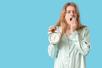 Young woman in pajamas with alarm clock suffering from insomnia on blue background