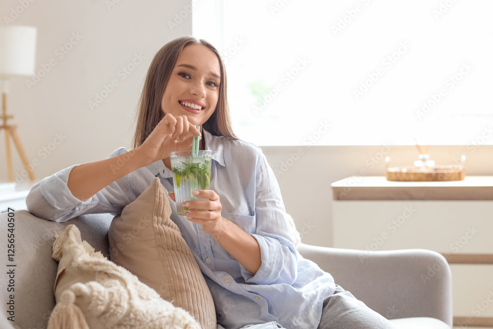 Poster Beautiful young happy woman with glass of mojito sitting on sofa at home